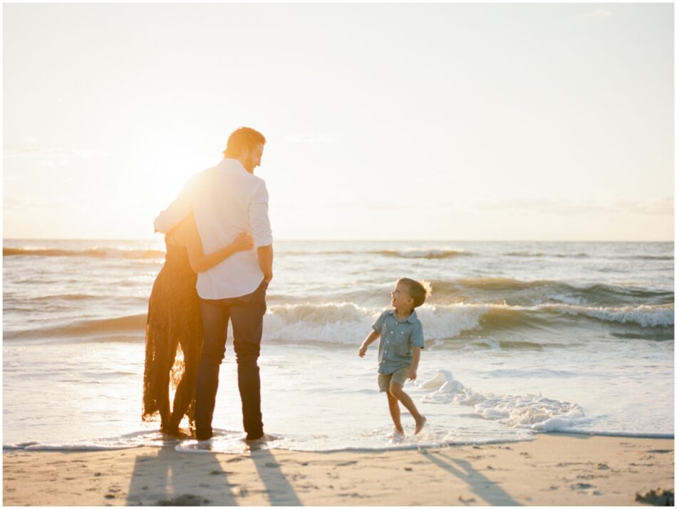 A family standing together at sunset for their family session in Naples, Florida. The mom and dad are holidng each other and the son is looking at them. The sun is setting over the water behind them