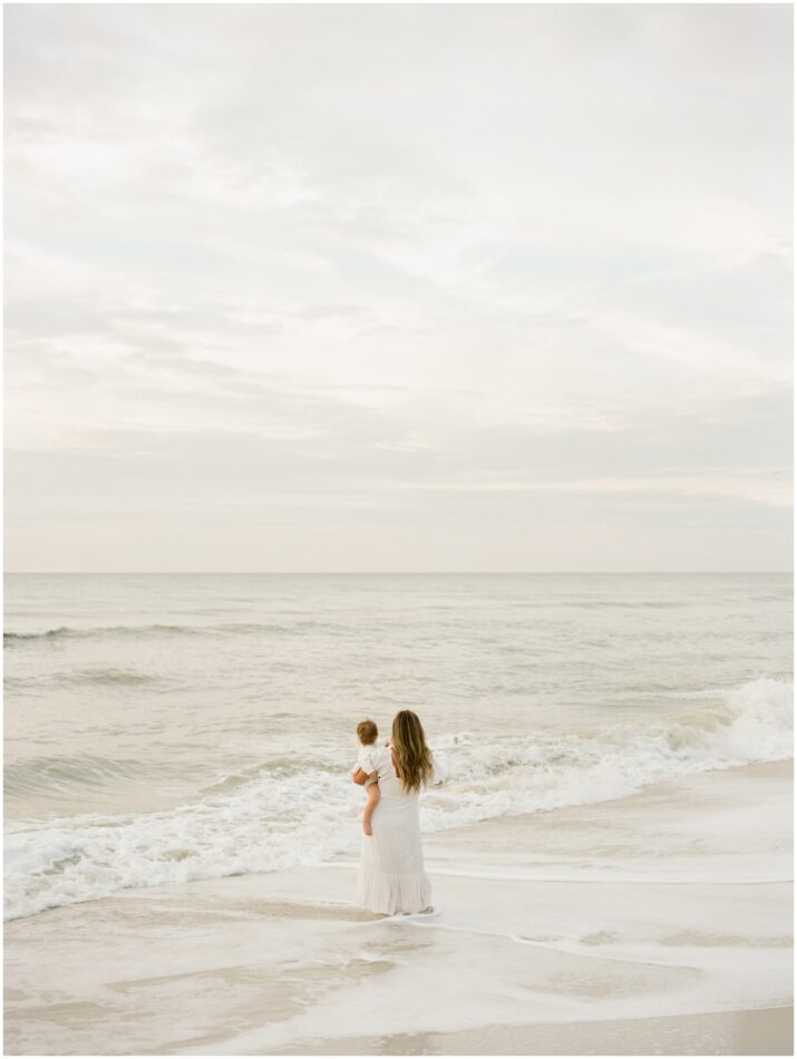 mother holding a baby facing the ocean behind her with waves crashing into the shore